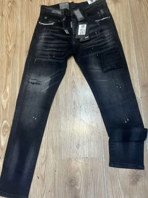Svarta jeans med slitningar och fickor - Snygga svarta jeans från Sixth June med slitningar, vita färgstänk och extra ficka på benet. Jeansen har en modern, smal passform och detaljer som dragkedjor och markerade sömmar. Perfekta för en trendig streetwear-look. passar till den som är 170-180cm vikt 70-80kg pris kan diskuteras helt oanvänd