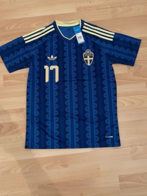 Sverige bortatröja Gyökeres #17 Adidas - Blå bortatröja för Sveriges landslag med Gyökeres och nummer 17 i gult. Tröjan har mönstrade blå ränder, gula axelränder och broderat landslagsmärke. Tillverkad i lätt och ventilerande material, perfekt för fotbollsträning eller match.