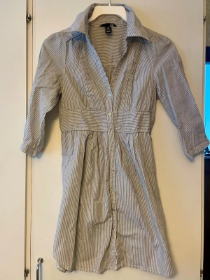 Randig skjortklänning från H&M i stl 34 - Snygg skjortklänning från H&M med smala vita och grå ränder. Klänningen har trekvartsärm, knappar hela vägen framtill och markerad midja. Klassisk krage och lätt, luftig bomullskvalitet gör den perfekt för varmare dagar. Normal i stl.  Använd fåtal ggr, inget att anmärka på.