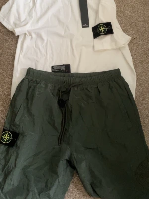 Stone Island set med shorts och t-shirt - Snyggt set från Stone Island med vita t-shirten och gröna shorts. Båda plaggen har den ikoniska Stone Island-loggan på ärm och ben. T-shirten är enkel och stilren, shortsen har resår i midjan och praktiska fickor. Perfekt för en avslappnad och trendig look.