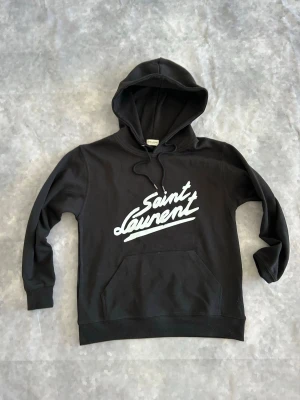 Svart Saint Laurent hoodie - Svart hoodie från Saint Laurent med vit logga tryckt på bröstet. Klassisk modell med huva och dragsnören samt stor magficka framtill. Tillverkad i mjukt bomullsmaterial som är skönt att bära. Perfekt för en avslappnad och trendig stil.