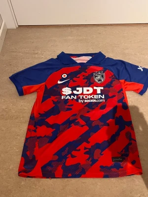 JDT matchtröja röd/blå Nike - Snygg JDT matchtröja från Nike i rött och blått med kamouflagemönster. Tröjan har klubbmärke och sponsortryck på bröstet samt klassisk krage. Perfekt för fotbollsträning eller att bära som supporter.