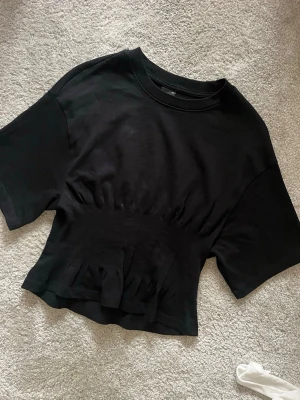 Svart topp - Säljer en svart croppad topp med rynkade detaljer i midjan och ärmar till armbågarna. Toppen har en rund halsringning. Perfekt för en trendig och avslappnad look.