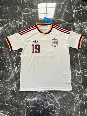 Vit Spanien tröja Adidas retro - Retroinspirerad vit Spanien-tröja från Adidas med vinröda och gula detaljer. Tröjan har nummer 19 på bröstet, Adidas-logga och Spaniens landslagsemblem. Klassiska tre ränder på axlarna och V-ringad krage i vinrött och gult. Tillverkad i lätt, funktionellt material.