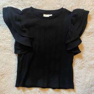 Svart volangblus från Gina Tricot - Supersnygg svart blus från Gina Tricot med volangdetaljer på axlarna och ribbad struktur.  Materialet är mjukt och stretchigt, vilket gör den bekväm att bära hela dagen. Använd 1-2 gånger❣️❣️