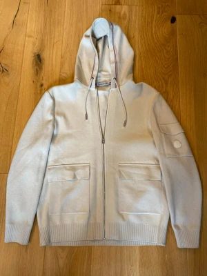 Beige kofta med dragkedja från Moncler - Snygg beige kofta från Moncler med dragkedja framtill, två stora fickor och en extra ficka på ärmen. Tröjan har ribbade muddar och huva med dragsko. Perfekt för en avslappnad och stilren look.