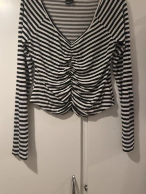 Randig långärmad topp Gina Tricot - Snygg långärmad topp från Gina Tricot med svartvita ränder och v-ringning. Toppen har rynkade detaljer framtill som ger en cool look. Perfekt att styla med jeans eller kjol för en trendig outfit.