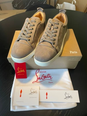 Gråa Christian Louboutin sneakers - Snygga gråa sneakers från Christian Louboutin i mocka med klassisk röd sula. Skorna har lågt skaft, rund tå och vita gummisulor. Kommer med originalkartong, dustbag och certifikat. Perfekta för dig som vill sticka ut med exklusiva detaljer.