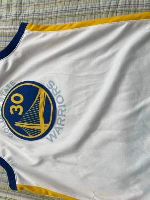 Golden State Warriors basketlinne - Säljer ett vitt Golden State Warriors basketlinne med blå och gula detaljer. Framsidan har lagets logga och nummer 30, baksidan har stort nummer 30 och texten Curry. Perfekt för basketträning eller att bära som supporter.