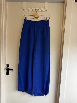 Blå vida chinos från Monki - Stilrena blå chinos från Monki i storlek 36. Byxorna har hög midja, vida ben och klassisk knappstängning. Materialet är mjukt och ger en skön känsla. Perfekta för dig som gillar en trendig och avslappnad look. Fållsömmen på vänster ben har släppt, men använd endast fåtal gånger.