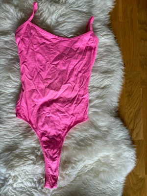 Rosa ribbad body med smala axelband - Snygg rosa body i ribbat material med smala axelband och hög skärning. Bodyn har en djup rygg och är stretchig för en bekväm passform. Perfekt att styla till jeans eller kjol för en trendig look.