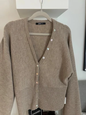 Beige kofta från Gina Tricot - Beige stickad kofta från Gina Tricot med v-ringning och glansiga knappar framtill. Koftan har långa ärmar och bred ribbstickad nederkant som ger en snygg siluett. Perfekt att bära över en topp eller t-shirt.