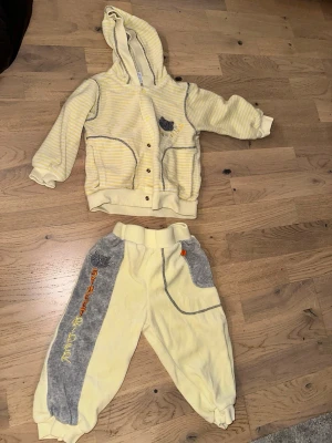 Gult och grått mjukisset med huva - Gult och grått mjukisset med huva Mysigt tvådelat set i mjuk frotté med gul bas och grå detaljer. Jackan har huva, knappar och två fickor samt ett litet broderat kattmotiv. Byxorna har resår i midjan och grå paneler med broderad text. Perfekt för chill dagar hemma.
