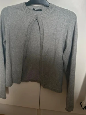 Grå kofta från Gina Tricot - En stilren grå kofta från Gina Tricot med rund halsringning och knapp framtill. Koftan har långa ärmar och ribbade muddar vid ärmslut och nederkant. Perfekt att slänga över en t-shirt för en chill look.