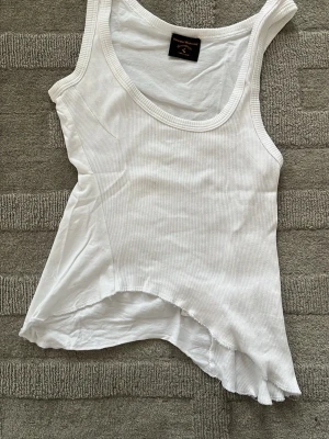 Vit ribbad linnetopp från Vivienne Westwood - Säljer en vit ribbad linnetopp från Vivienne Westwood med asymmetrisk nederkant och breda axelband. Toppen har en cool, lite edgy design och är gjord i mjuk bomull. Perfekt att styla med jeans eller kjol för en trendig look.