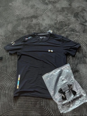 Svart träningströja från Under Armour - Svart kortärmad träningströja från Under Armour med logga på bröstet och färgglada detaljer på ärm och sida. Tillverkad i ett lätt och snabbtorkande material som passar perfekt för intensiva träningspass.