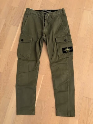 Olivgröna cargobyxor från Stone Island - ÄKTA!! Snygga olivgröna cargobyxor från Stone Island med klassisk patch på benet. Byxorna har flera fickor med lock och knappstängning samt smal passform. Perfekta för en streetwear-look och tillverkade i slitstark bomull. Måtten finns bland dem sista bilderna. 