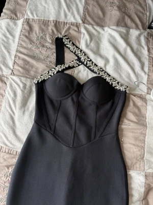 Svart långklänning med pärldetaljer - Elegant svart långklänning med figurnära passform och unik oneshoulder-design. Klänningen har en axelrem dekorerad med pärlor och en markerad byst med kupor. Perfekt för dig som vill sticka ut med en stilren och lyxig look.