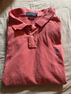 Rosa pikétröja från Polo Ralph Lauren - Klassisk rosa pikétröja från Polo Ralph Lauren med korta ärmar och krage med knappar. Tröjan har broderad logga på bröstet och är tillverkad i bomull. Perfekt för en avslappnad och stilren look.