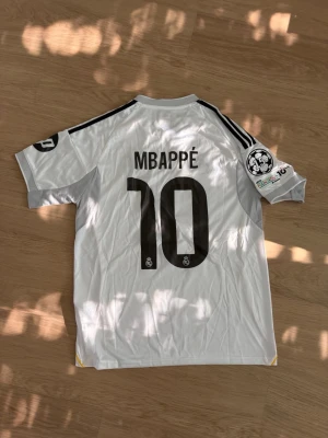 Vit Real Madrid Mbappé tröja - Säljer en vit Real Madrid matchtröja med Mbappé och nummer 10 på ryggen. Tröjan har svarta detaljer, Champions League-märke på ärmen och klassiska Adidas-ränder på axlarna. Perfekt för fotbollsfans som vill sticka ut på planen eller läktaren.