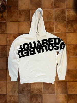 Vit hoodie från DSQUARED2 XL - Snygg vit hoodie från DSQUARED2 i storlek XL med stor svart logga tryckt tvärs över bröstet. Klassisk känguruficka framtill, dragsko i huvan och ribbade muddar. Perfekt för dig som gillar streetwear och vill sticka ut.