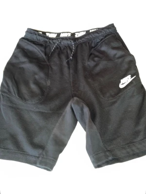 Svarta Nike shorts med snörning - Svarta sportiga shorts från Nike med vit logga på vänster ben. Shortsen har snörning i midjan och är tillverkade i ett mjukt och lätt material som passar perfekt för träning eller chill. Två stora fickor framtill och elastisk midja för skön passform.