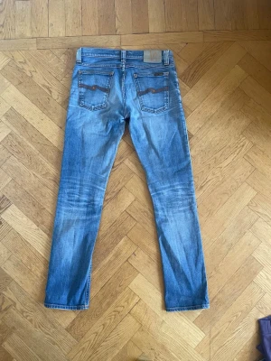 Blå slim jeans från Nudie Jeans - Säljer ett par klassiska blå slim jeans från Nudie Jeans med snygga slitningar och kontrastsömmar. Jeansen har fem fickor och en normal midja. Perfekta för dig som gillar en tidlös och avslappnad stil.
