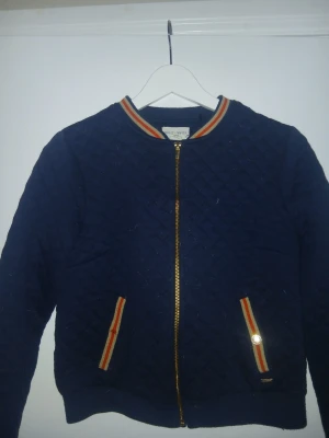 Mörkblå quiltad bomberjacka - Quiltad mörkblå bomberjacka från Holly & White med guldfärgad dragkedja och ribbade muddar. Krage och fickor har ränder i orange, vitt och gult för en sportig look. Jackan har en klassisk bomberpassform och är tillverkad i mjukt tyg. En knapp på höger fick saknas vilket man kan se på bild nummer 1