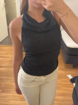 Svart halterneck topp med drapering - Snygg svart halterneck topp med draperad halsringning. Toppen är ärmlös och har en tight passform som framhäver figuren. Perfekt att styla med jeans eller kjol för en trendig look.