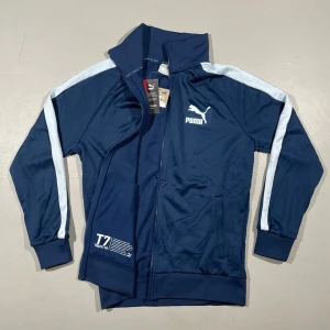 Marinblå Puma T7 track jacket - Snygg marinblå track jacket från Puma med ljusaste av blåa ränder längs ärmarna. Den klassiska T7 Track 1968-modellen. Jackan har dragkedja, ståkrage och ribbade muddar. Nypris i butik (för samma skick som hos oss) 899:-