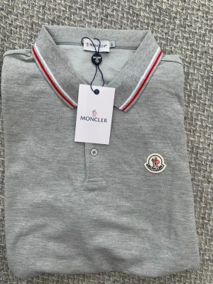Grå pikétröja från Moncler - Snygg grå pikétröja från Moncler med klassisk krage och knäppning framtill. Kragen har röda och vita ränder och bröstet pryds av Monclers ikoniska logotyp. Perfekt för en stilren och sportig look.