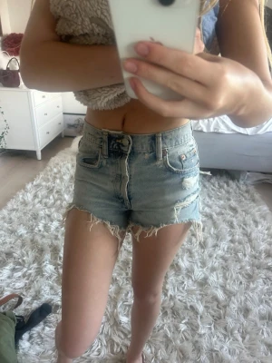 Ljusblå jeansshorts med fransig kant - Säljer ett par ljusblå denimshorts med hög midja och fransig, rå kant. Klassisk femficksmodell med dragkedja och knapp framtill. Perfekta till sommaren och lätta att matcha med allt.