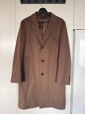 Brun rutig ullkappa med knappar - Stilren brun kappa i rutigt mönster med tre knappar framtill från Uniqlo. Kappan har två snedställda fickor och är helfodrad med slät insida. Storlek: Herr 46 (Small)