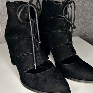 Snygga Cut-Out Ankelboots i Mocka – Stl 37 - Säljer ett par supersnygga svarta klackskor / ankelboots i fin mockaimitation i storlek 37 (art.nr 35-49074). Riktigt snygg design med cut-out detaljer på sidorna, spetsig tå och dekorativ snörning. Smidiga YKK-dragkedjor i hälen så de är enkla att ta på och av! Stadig blockklack med en klackhöjd på 9 cm. Absolut toppskick, knappt använda vilket syns på de helt felfria sulorna (se bilder). Perfekta till festen! Kan skickas säkert med Plicks frakt.