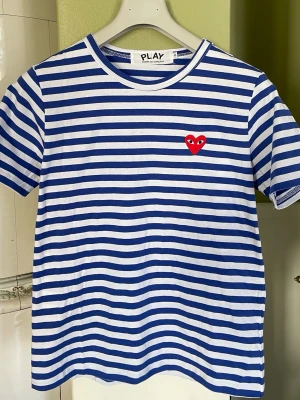 Randig t-shirt från Comme des Garçons - Säljer en blåvit randig t-shirt från Comme des Garçons Play med det ikoniska röda hjärtat broderat på bröstet. T-shirten har rund hals, korta ärmar och är gjord i mjuk bomull. Perfekt för dig som gillar streetstyle och vill sticka ut lite extra.