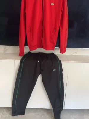 Röd och svart träningsdress Lacoste - Säljer en snygg träningsdress från Lacoste med röd zip-jacka och svarta byxor. Jackan har gröna detaljer längs ärmarna och dragkedja framtill, medan byxorna har gröna linjer längs sidorna och resår i midjan. Båda delarna har Lacoste-loggan broderad. Storlek S skickas samma vardag 