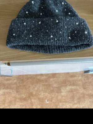 Mörkgrå stickad mössa med pärlor - Snygg mörkgrå stickad mössa dekorerad med små vita pärlor över hela ytan. Mössan har en bred uppvikt kant och är gjord i ett mjukt, värmande material som passar perfekt för kyliga dagar. Stilren och trendig accessoar för höst och vinter.