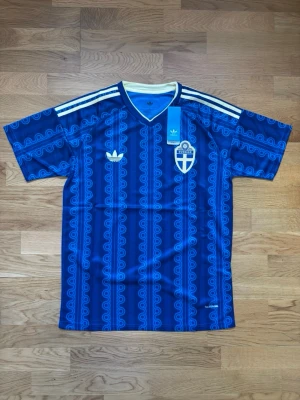 Blå Sverige vm 2026 fotbollströja  - Retroinspirerad blå fotbollströja från Adidas med Sveriges landslagsemblem på bröstet. Tröjan har ett unikt mönster, vita Adidas-ränder på axlarna och V-ringad krage med ljusgul kant. Perfekt för dig som vill sticka ut på läktaren eller träningen.