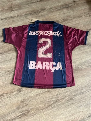 Barça x Cactus Jack matchtröja - Säljer en limited edition Barcelona x Cactus Jack matchtröja i vinrött och marinblått med breda ränder. Tröjan har tryck med nummer 2 och texten BARÇA på ryggen samt klubbmärke och Nike-logga på bröstet. Tillverkad i lätt och ventilerande material, perfekt för fotboll eller streetwear.