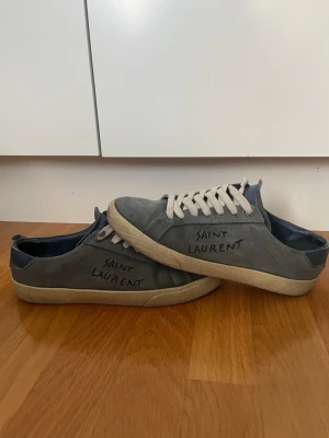 Grå Saint Laurent sneakers i mocka - Grå sneakers från Saint Laurent i mjuk mocka med vita snören och beige sula. Sidan pryds av handskriven Saint Laurent-logga. Klassisk låg modell med rund tå och diskreta detaljer. Perfekta för en avslappnad och trendig stil.