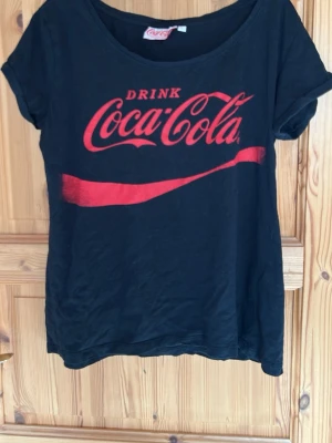 Svart Coca-Cola t-shirt med röd logga - Cool svart t-shirt med stor röd Coca-Cola-logga och texten 'Drink Coca-Cola' på bröstet. Klassisk rund halsringning och korta ärmar. Perfekt för dig som gillar retrostil och vill sticka ut lite extra.