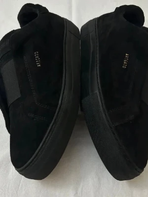 Svarta sneakers från Axel Arigato - Stilrena svarta sneakers från Axel Arigato i mocka med diskret logga i guld på sidan och plösen. Skorna har en låg silhuett, svart sula och elastiskt band över vristen för en modern look. Perfekta för dig som gillar minimalistisk och trendig stil.