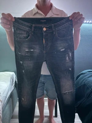 Svarta slitna jeans från Alessandro - Säljer ett par mörkblåa jeans från Alessandro med slitningar och distressed detaljer framtill. Jeansen har fem fickor, knappgylf och smal passform. Perfekta för dig som gillar en edgy och trendig stil.