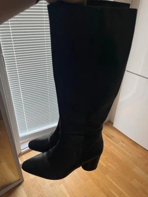 Svarta knähöga boots med klack - Snygga svarta knähöga stövlar i slätt material med bred klack och spetsig tå. Använda fåtal gånger, köpta för 1365 kr. 
