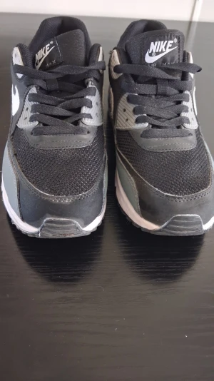 Nike Air Max 90 svart/grå sneakers - Nike Air Max 90 sneakers i svart och grått med vita detaljer. Ovandelen är i mesh och syntetmaterial för bra andningsförmåga och komfort. Klassisk Air Max-dämpning i sulan och vit yttersula. Swoosh-logga på sidan och Air Max-märke på plösen.