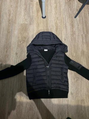 Moncler cardigan - Snygg svart dunväst med vadderad huva och dragkedja framtill. Västen har ribbade detaljer vid midjan och två fickor med dragkedja. Perfekt för lager-på-lager och streetwear-vibe. Passar till både hoodie och t-shirt.