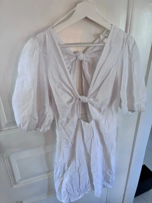 Vit kort klänning från H&M - Säljer en vit kort klänning från H&M med puffärmar och två knyt framtill. Klänningen har djup v-ringning och är tillverkad i ett lätt bomullstyg. Perfekt för varma dagar och har en trendig, luftig passform.