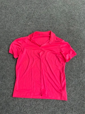 Adidas Rosa sportpiké med kort ärm - Snygg rosa sportpiké med korta ärmar och klassisk krage. Tillverkad i ett lätt och snabbtorkande material som passar perfekt för träning eller golf. Enkel design. 