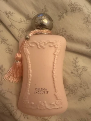 Delina Exclusif parfymflaska - Elegant parfymflaska i matt ljusrosa med dekorativa reliefdetaljer och en silverfärgad rund kork. Flaskan har en rosa tofs som dekoration och texten 'Delina Exclusif' framtill. Märket Parfums de Marly är ingraverat på baksidan. Det finns cirka 80% kvar av flaskan om inte ens mer flaskan är 75 ml stor 