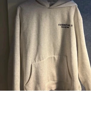 Essentials Fear of God beige hoodie - Beige hoodie från Essentials Fear of God med svart logga på bröstet och ryggen. Klassisk känguruficka framtill och huva. Perfekt för en avslappnad och trendig streetwear-look.